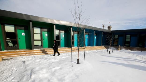 A woman walks through snow next to a black building fro<em></em>nted by blue and green doors next to windows.