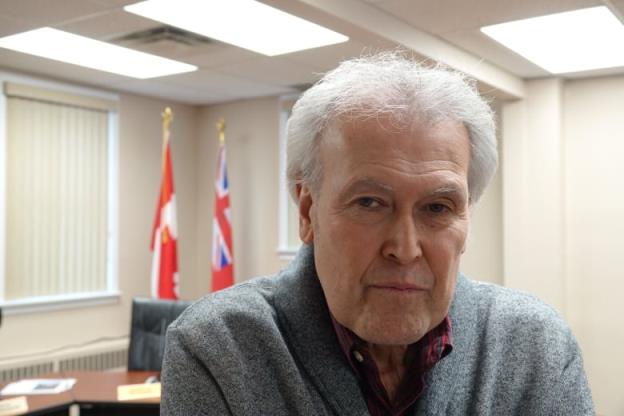 A man with white hair stares at the camera with a serious face. The flags of o<em></em>ntario and Canada can be seen in the background behind him.