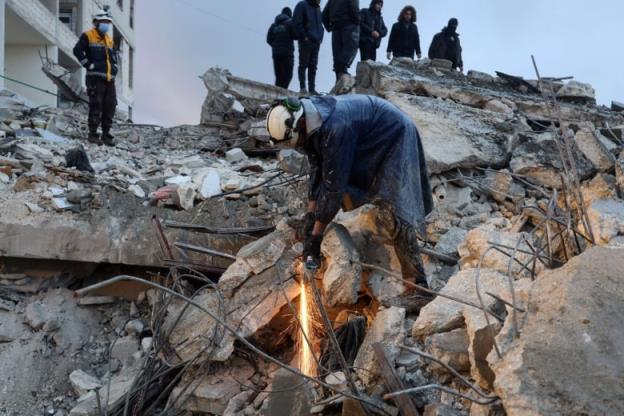 A man with a white helmet cuts through co<em></em>ncrete as other people stand above, on a pile of rubble, watching. 