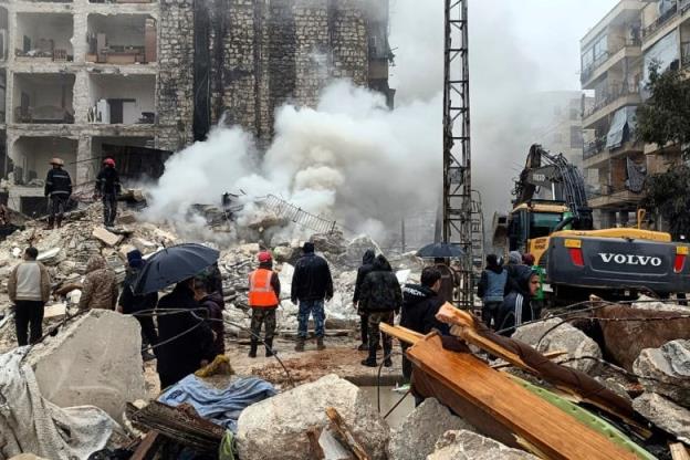 A cloud rises from a pile of co<em></em>ncrete rubble as people stand in front. 